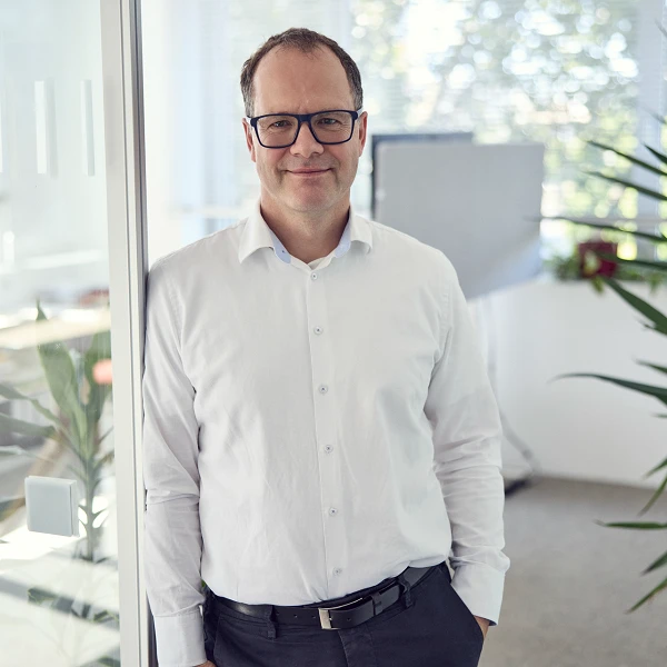 Ein Mann in einem weißen Hemd und Brille steht in einem modernen Büro mit großen Fenstern und Pflanzen im Hintergrund.