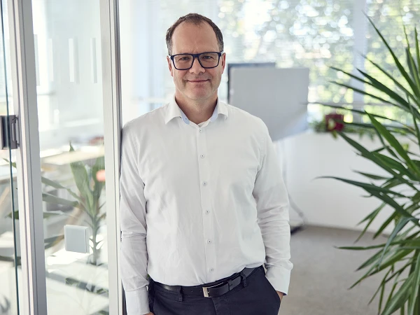 Ein Mann in einem weißen Hemd und Brille steht in einem modernen Büro mit großen Fenstern und Pflanzen im Hintergrund.