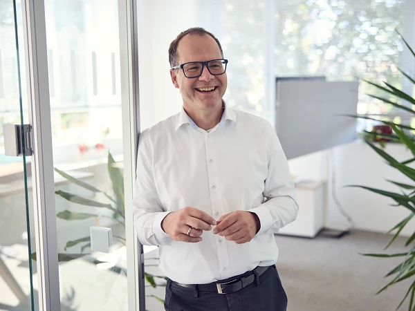 Ein Mann mit Brille und einem weißen Hemd lächelt in einem hellen Büro, umgeben von Pflanzen und natürlichem Licht.