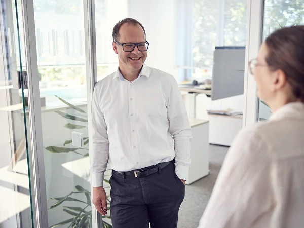 Ein Mann in einem weißen Hemd und einer Brille lächelt, während er mit einer Frau spricht, die ihm gegenübersteht. Die Szene spielt sich in einem modernen Büro mit Glaswänden und Pflanzen im Hintergrund ab.