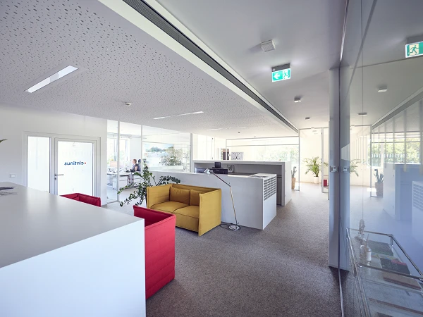 Modernes Büro mit offenen Räumen, einer Empfangstheke und bequemen Sofas in Gelb und Rot, sowie Pflanzen und großen Fenstern, die viel Tageslicht hereinlassen.