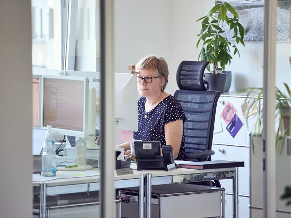 Eine Frau mit kurzer, blonder Frisur und Brille sitzt an einem Schreibtisch in einem modernen Büro, konzentriert auf ihre Computermonitore. Im Hintergrund sind Pflanzen und Schreibtischutensilien sichtbar.