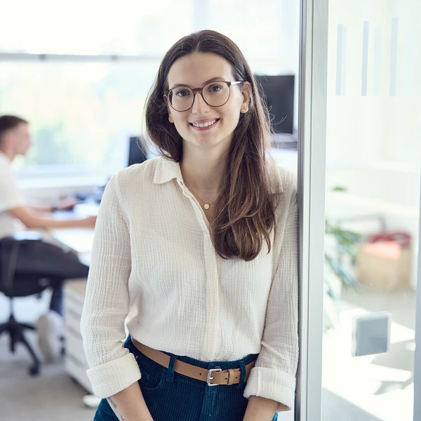 Eine junge Frau mit langen, braunen Haaren und einer blauen Jeans steht lächelnd neben einer Glaswand in einem modernen Büro, während sich im Hintergrund ein Kollege am Schreibtisch mit einem Computer beschäftigt.