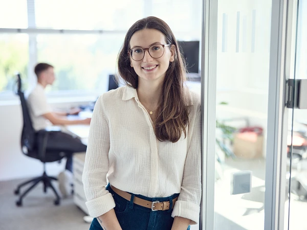 Eine junge Frau mit langen, braunen Haaren und einer blauen Jeans steht lächelnd neben einer Glaswand in einem modernen Büro, während sich im Hintergrund ein Kollege am Schreibtisch mit einem Computer beschäftigt.