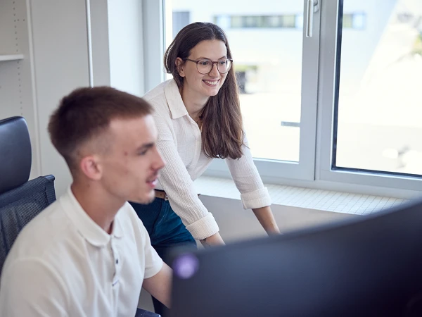Eine Frau mit langen, braunen Haaren und Brille steht lächelnd hinter einem Mann mit kurzem, blondem Haar, der am Computer sitzt. Im Hintergrund sind Fenster mit natürlichem Licht und ein moderner Bürobereich sichtbar.