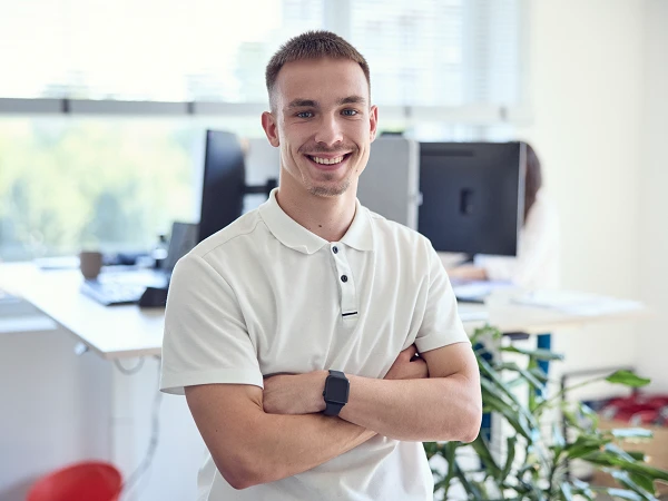 Ein junger Mann mit einem breiten Lächeln steht lässig in einem modernen Büro, während er die Arme vor der Brust verschränkt. Im Hintergrund sind Schreibtische und Computer zu sehen.