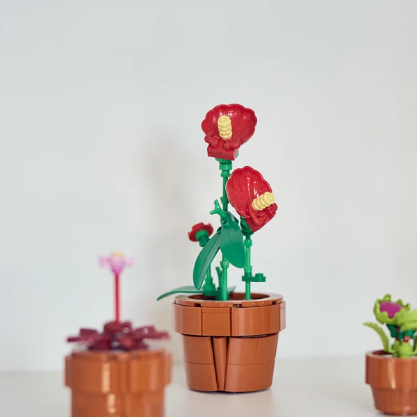 Bunte Pflanzendekorationen aus Kunststoff in Tontöpfen auf einem Regal, darunter eine kleine pinkfarbene Blume, eine grüne Pflanze mit roten Blüten und zwei grüne Figuren, die wie Pflanzen aussehen.