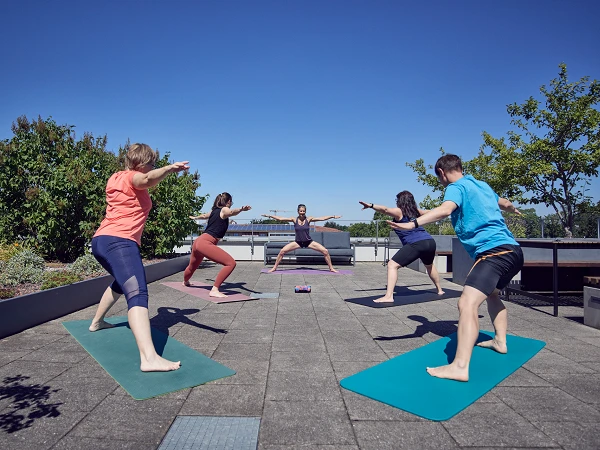 Gruppentraining im Freien auf einer Dachterrasse, bei dem fünf Personen unterschiedliche Yoga-Posen in einem sonnigen Umfeld einnehmen. Im Hintergrund sind Bäume und der blaue Himmel sichtbar.