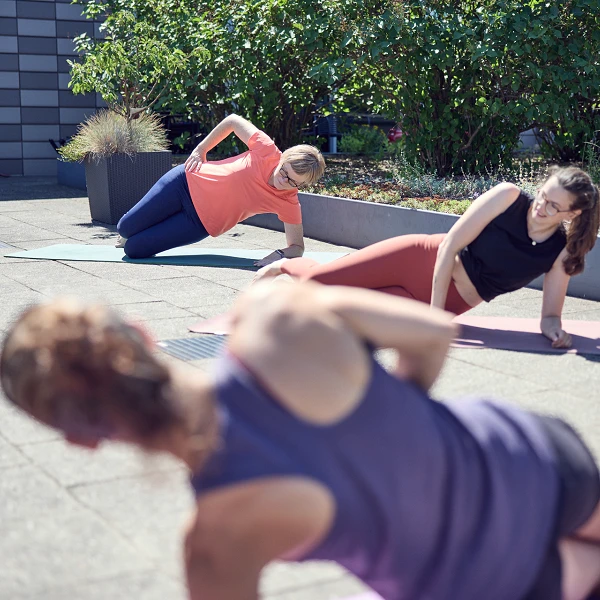 Drei Frauen üben auf Yogamatten im Freien, während sie seitliche Plankenhaltungen einnehmen. Man sieht eine grüne Bepflanzung im Hintergrund und einen hellen, sonnigen Himmel.