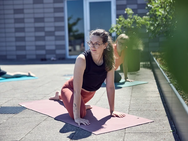 Eine Frau in Sportbekleidung übt eine Yoga-Position auf einer Matte im Freien, während im Hintergrund eine weitere Person ebenfalls Yoga praktiziert. Die Umgebung wirkt hell und freundlich, mit Pflanzen und moderner Architektur.