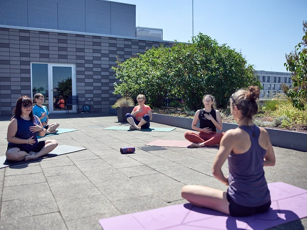 Sechs Personen sitzen auf Yogamatten auf einer Terrasse und praktizieren entspannt Yoga. Im Hintergrund sind grüne Pflanzen und moderne Gebäude sichtbar.