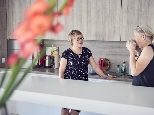 Zwei Frauen stehen in einer modernen Küche, eine von ihnen trägt ein schwarzes Oberteil mit weißen Punkten und lächelt, während die andere einen Becher hält und in eine nachdenkliche Pose versunken ist. Im Vordergrund sind unscharf Blumen zu sehen.