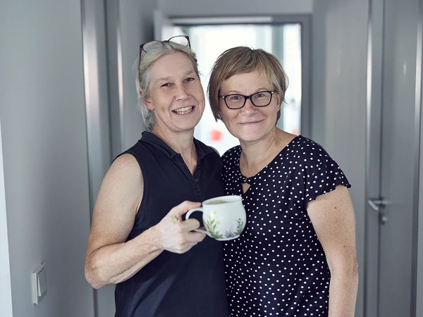 Zwei Frauen stehen in einem hellen Flur und lächeln in die Kamera, während eine von ihnen eine Tasse in der Hand hält. Beide tragen casual Kleidung, eine ist in einem ärmellosen Oberteil und die andere in einem gepunkteten Shirt gekleidet.