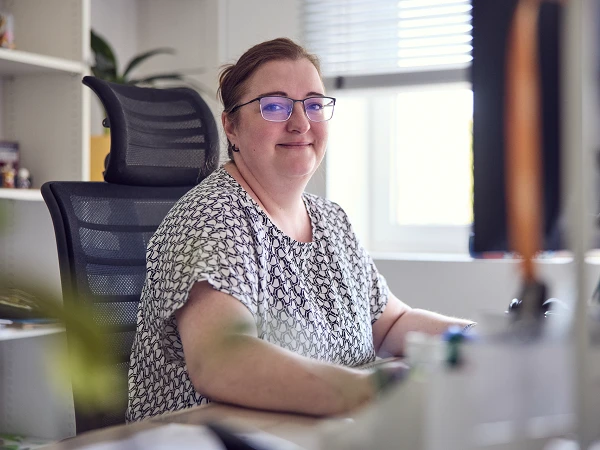 Eine lächelnde Frau mit Brille sitzt an einem Schreibtisch in einem hellen Büro. Im Hintergrund sind Pflanzen und Regale sichtbar.