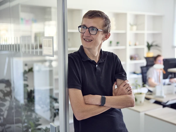 Ein junger Mann mit Brille und kurzen Haaren steht lächelnd an einer Glaswand in einem modernen Büro, während im Hintergrund ein weiterer Kollege an einem Schreibtisch arbeitet.