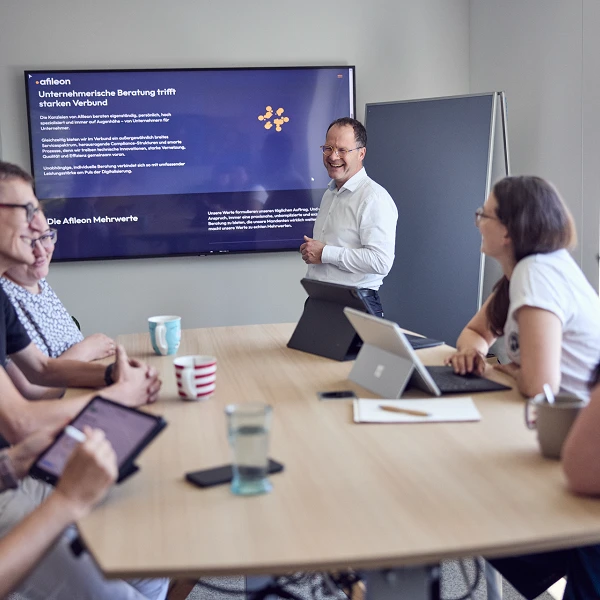 Ein Mann steht in einem Konferenzraum und präsentiert eine Folie auf einem Bildschirm, während sechs Personen am Tisch aufmerksam zuhören und auf Tablets sowie Laptops arbeiten.