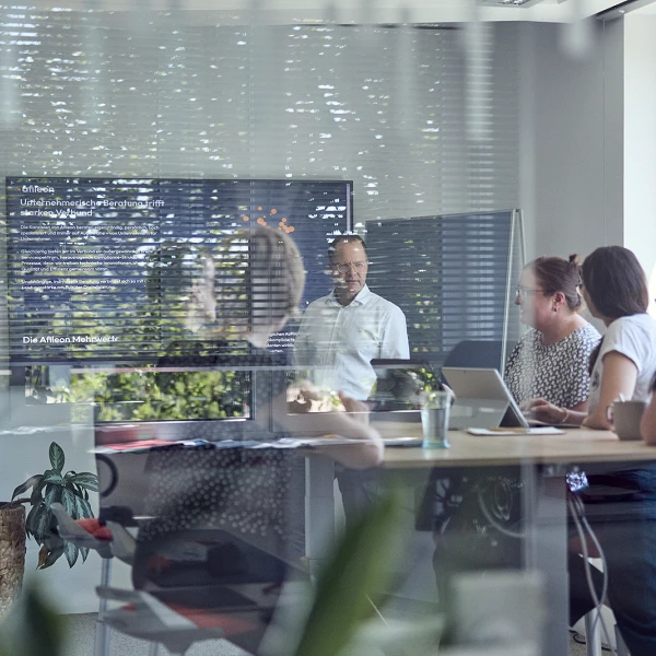 Eine Gruppe von Menschen sitzt an einem Tisch in einem modernen Konferenzraum, während ein Mann im Vordergrund eine Präsentation hält. Im Hintergrund sind Pflanzen und Fenster zu sehen, die viel Tageslicht hereinlassen.