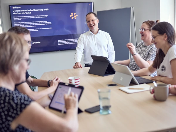 Eine Gruppe von fünf Personen sitzt an einem Tisch in einem Besprechungsraum, während ein Mann im Vordergrund, der lächelt, eine Präsentation leitet. Auf dem Tisch liegen Tablets und ein Glas Wasser, während im Hintergrund eine Präsentationswand zu sehen ist, die Informationen über Unternehmensberatung zeigt.