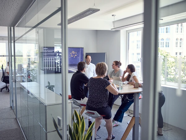 Eine Gruppe von fünf Personen sitzt an einem Stehtisch in einem modernen Büro und diskutiert; ein Mann steht und präsentiert Informationen auf einem Bildschirm im Hintergrund. Im Raum ist eine entspannte Arbeitsatmosphäre zu erkennen.