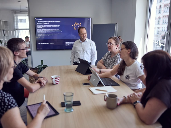 Eine Gruppe von sechs Personen sitzt an einem Tisch in einem modernen Besprechungsraum, während eine Präsentation im Hintergrund an einer Wand gezeigt wird. Einer der Teilnehmer, ein Mann in der Mitte, steht und spricht, während die anderen auf Tablets und Laptops schauen. Auf dem Tisch befinden sich verschiedene Getränke und Notizmaterialien.