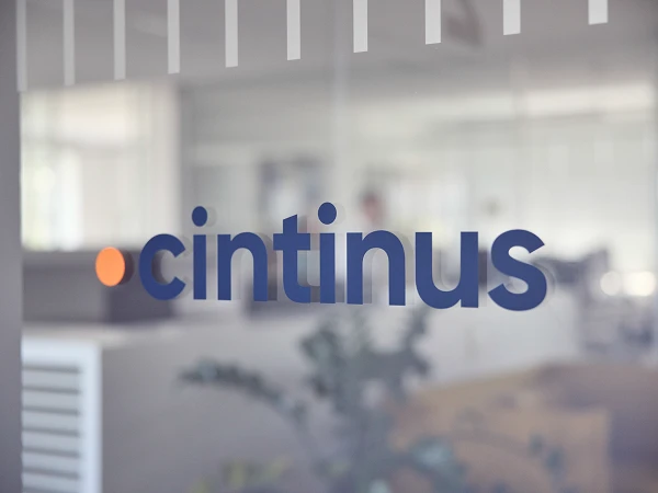 Das Bild zeigt das Logo von "cintinus", das auf einer Glaswand angebracht ist. Im Hintergrund ist ein helles, modernes Büro mit einigen Pflanzen sichtbar.