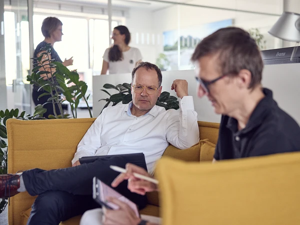 Zwei Männer sitzen auf einem gelben Sofa im Büro; einer mit einer Brille hält ein Tablet und hört aufmerksam zu, während der andere auf sein Smartphone schaut. Im Hintergrund sind weitere Personen zu sehen, die sich unterhalten.