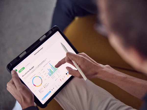 Eine Person zeigt mit einem Stift auf ein Tablet, das ein Diagramm zur Budgetverwaltung darstellt, während sie auf einer Couch sitzt.