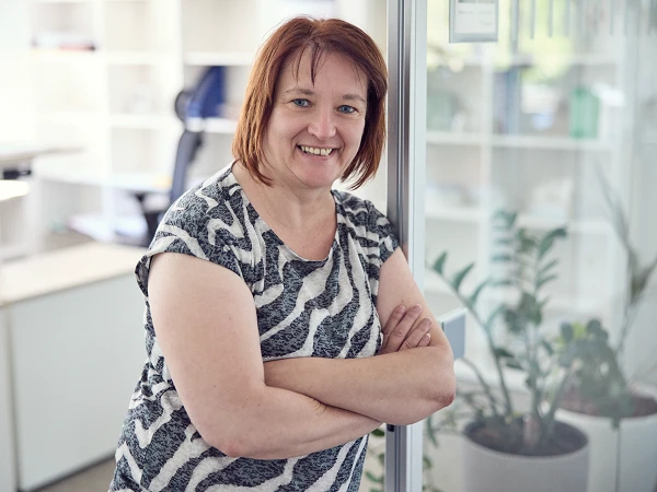 Eine lächelnde Frau mit kurzen, braunen Haaren in einem gestreiften Shirt steht mit verschränkten Armen in einem modernen Büro, das von Pflanzen und Regalen mit Büromaterialien umgeben ist.