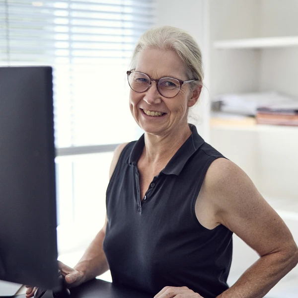 Eine älterer Frau mit Brille und einem schulterlangen, grauen Haarschnitt sitzt an einem Schreibtisch und lächelt, während sie auf einen Computerbildschirm schaut. Im Hintergrund sind Regale mit aufgerollten Papieren und einem kleinen Gerätes sichtbar.