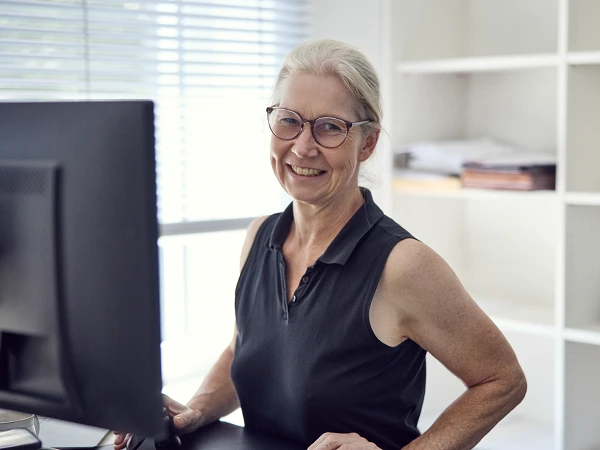 Eine älterer Frau mit Brille und einem schulterlangen, grauen Haarschnitt sitzt an einem Schreibtisch und lächelt, während sie auf einen Computerbildschirm schaut. Im Hintergrund sind Regale mit aufgerollten Papieren und einem kleinen Gerätes sichtbar.