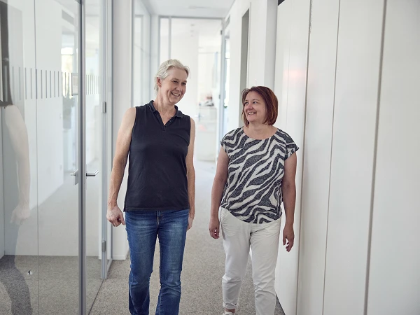 Zwei Frauen gehen lächelnd einen hellen Flur vorbei, der von Glaswänden und modernen Büromöbeln umgeben ist. Die ältere Frau trägt ein dunkles ärmelloses Oberteil und Jeans, während die andere Frau ein gestreiftes T-Shirt und helle Hosen trägt.