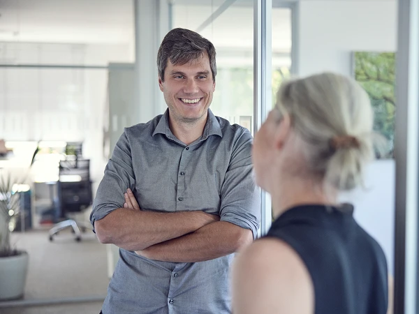 Ein Mann mit hellem Hemd steht in einem modernen Büro und lächelt, während er mit einer Frau spricht, die ihm den Rücken zugewandt hat. Im Hintergrund sind Büromöbel und Pflanzen sichtbar.