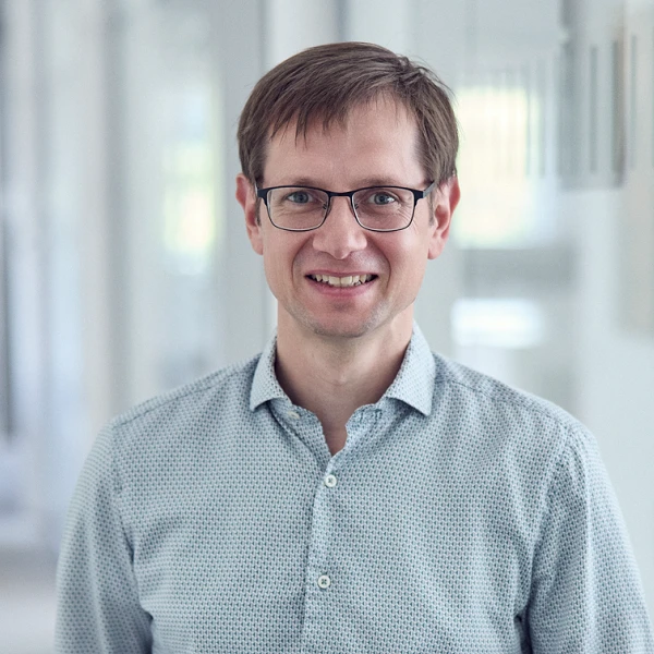 Ein Mann steht in einem modernen Büroflur, trägt ein kariertes Hemd und eine graue Hose und lächelt freundlich in die Kamera. Im Hintergrund sind Pflanzen und Glaswände sichtbar.