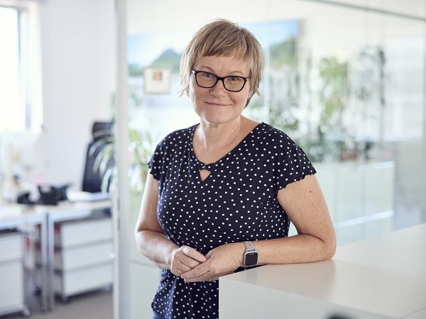 Eine Frau mit kurzen, strähnigen Haaren und Brille steht an einem Empfangstresen in einem modernen Büro, umgeben von Pflanzen und Büromöbeln, und lächelt in die Kamera. Sie trägt ein schwarzes Oberteil mit weißen Punkten.