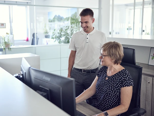 Ein Mann steht neben einer Frau, die an einem Computer sitzt. Beide schauen auf den Bildschirm, während es in einem modernen Büro mit vielen Pflanzen im Hintergrund hell und freundlich aussieht.
