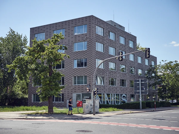 Ein modernes, vierstöckiges Gebäude mit einer grauen Klinkerfassade steht an einer Straßenecke. Vor dem Gebäude befindet sich ein kleiner Baum und ein Postkasten. An der Straßenecke steht eine rote Ampel. Die Umgebung ist grün und lichtdurchflutet, unter blauem Himmel.