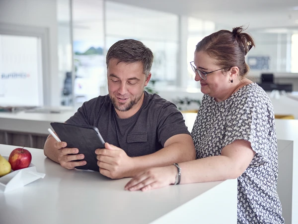 Ein Mann und eine Frau stehen an einem weißen Tisch und schauen gemeinsam auf ein Tablet. Der Mann lächelt, während die Frau interessiert zusieht. Im Hintergrund sind hell beleuchtete Büroräume zu erkennen, und auf dem Tisch liegen Äpfel.