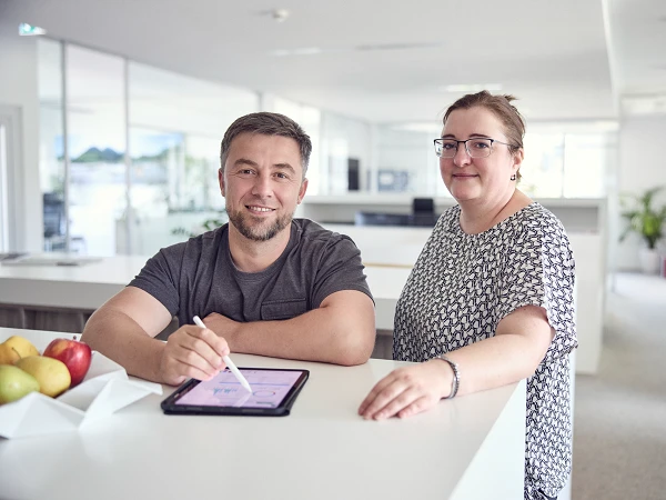 Ein Mann und eine Frau stehen an einem Empfangstresen in einem modernen Büro. Der Mann hält ein Tablet und zeigt darauf, während die Frau lächelt und ihre Hände auf dem Tresen hat. Im Hintergrund sind Fenster, Pflanzen und Büromöbel sichtbar.