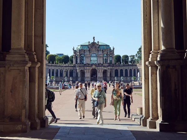 Blick durch einen eleganten Bogen auf eine belebte, sonnige Landschaft mit Menschen, die sich in der Ferne vor einem architektonisch ansprechenden Gebäude mit einem grünen Dach und einer Statue versammeln.
