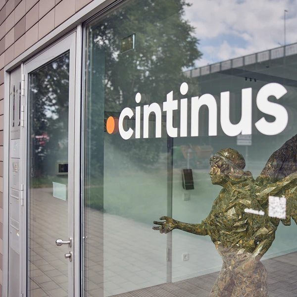Die Eingangstür eines Gebäudes mit dem Schriftzug "cintinus" und einer Skulptur eines Engels aus goldenen und grünen Materialien, die aus dem Fenster herausragt.