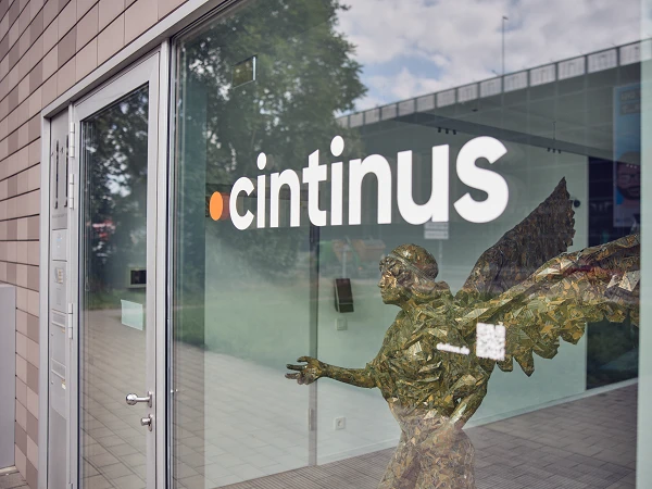 Die Eingangstür eines Gebäudes mit dem Schriftzug "cintinus" und einer Skulptur eines Engels aus goldenen und grünen Materialien, die aus dem Fenster herausragt.