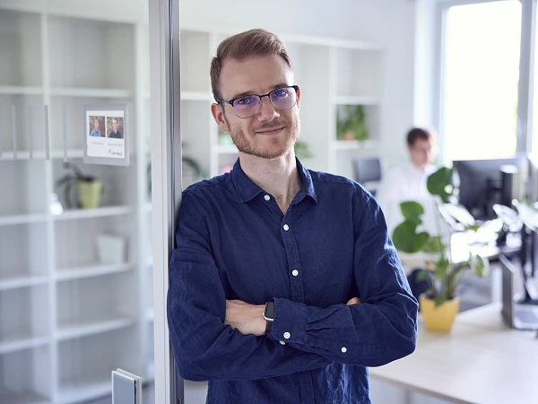Ein lächelnder Mann mit Brille steht mit verschränkten Armen neben einer Glaswand in einem modernen Büro, während im Hintergrund ein Kollege an einem Computer arbeitet.