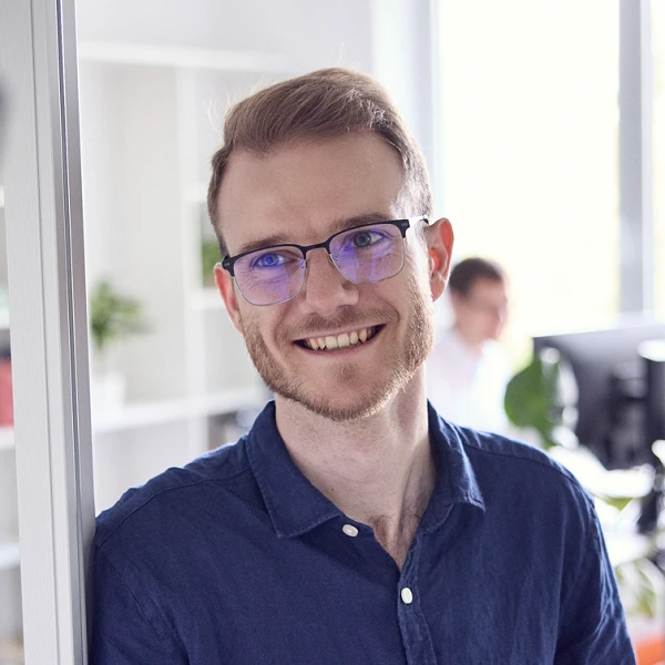 Zwei Männer in einem Büro unterhalten sich, während einer von ihnen lächelt und der andere am Fenster steht. Im Hintergrund sind Schreibtische mit Computern zu sehen.