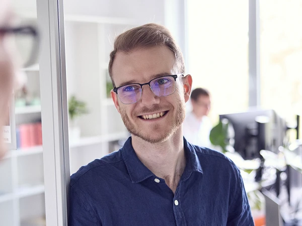 Zwei Männer in einem Büro unterhalten sich, während einer von ihnen lächelt und der andere am Fenster steht. Im Hintergrund sind Schreibtische mit Computern zu sehen.
