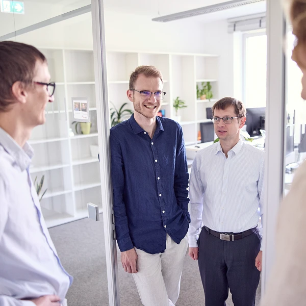 Vier Männer stehen in einem modernen Büro und unterhalten sich. Zwei der Männer tragen Brillen und lächeln, während der dritte Mann in einem hellblauen Hemd etwas schüchterner wirkt. Im Hintergrund sind Regale mit Pflanzen und Möbeln zu sehen.