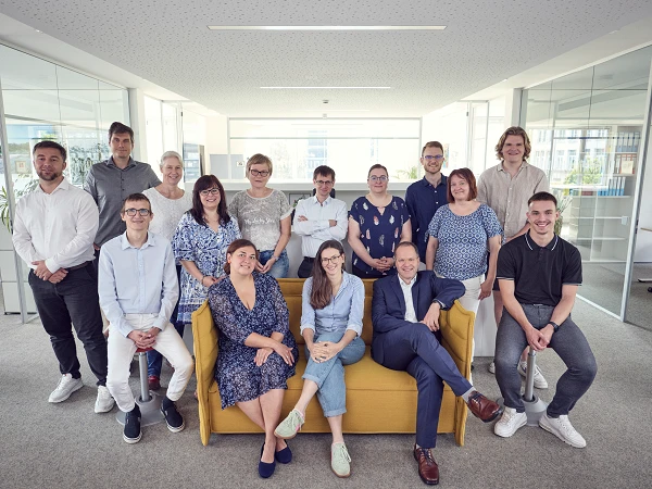 Eine Gruppe von 16 Personen sitzt und steht in einem modernen Büro mit großen Fenstern, das Tageslicht hereinlässt. Die meisten Personen lächeln und tragen legere Kleidung. Einige stehen im Hintergrund, während andere auf einem gelben Sofa sitzen.