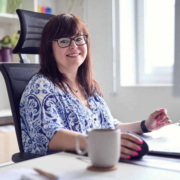 Eine lächelnde Frau mit Brille sitzt an einem Schreibtisch in einem hellen Büro und arbeitet am Computer, während sie eine Tasse in der Hand hält. Im Hintergrund sind Regale mit Pflanzen und Büroartikeln sichtbar.