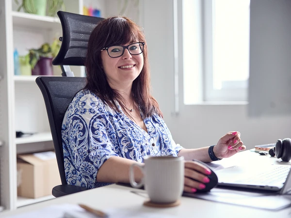Eine lächelnde Frau mit Brille sitzt an einem Schreibtisch in einem hellen Büro und arbeitet am Computer, während sie eine Tasse in der Hand hält. Im Hintergrund sind Regale mit Pflanzen und Büroartikeln sichtbar.
