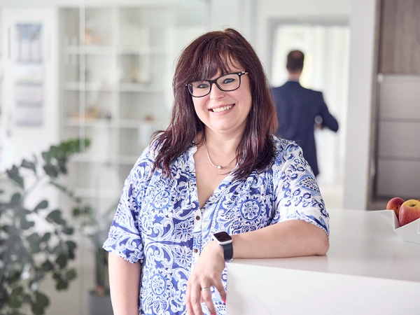 Eine lächelnde Frau mit langen, lockigen, brünetten Haaren und Brille steht an einem weißen Tisch, während sie in einem modernen, hellen Büro mit Pflanzen und einem verschwommenen Hintergrund联系客服.