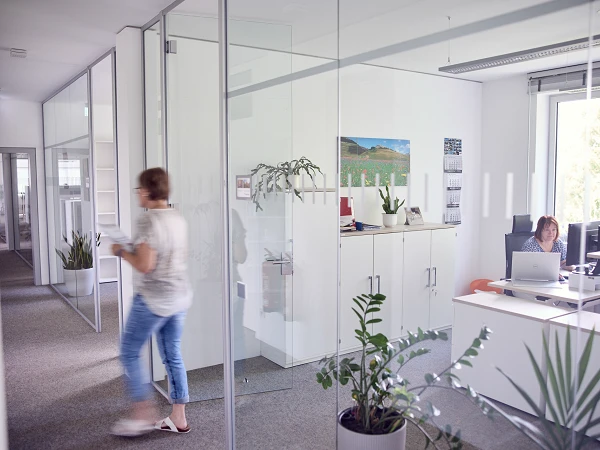 Eine Person geht durch ein modernes Büro mit Glaswänden, während eine andere Person an einem Schreibtisch mit Computer sitzt. Im Hintergrund sind Pflanzen und ein Bild an der Wand zu sehen.